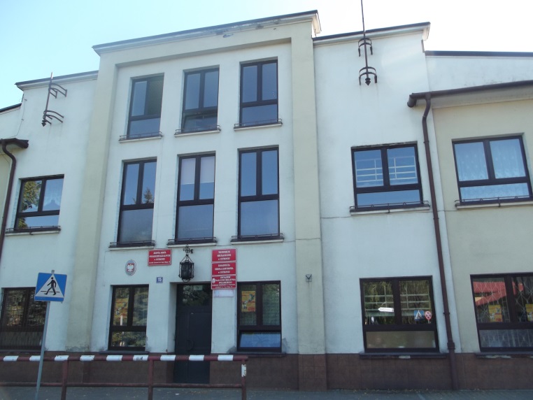 Trzykondygnacyjny budynek o jasnej elewacji z ciemnymi ramami okien. Centralnie umieszczone wejście z tablicami informacyjnymi. Przed budynkiem znak przejścia dla pieszych i barierka ochronna.